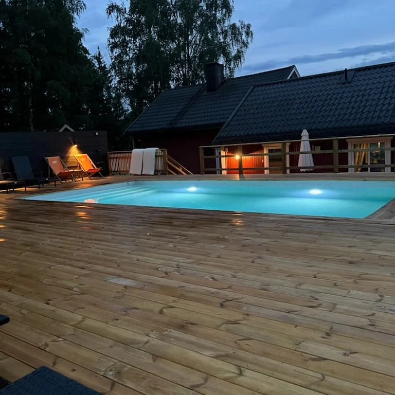 Trädäck runt pool vid skymning med rött trähus i bakgrunden